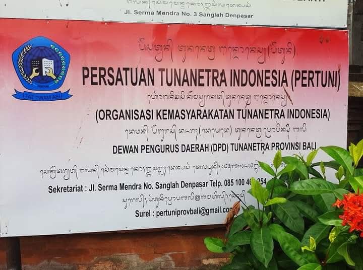 Terlihat sebuah papan nama organisasi yang terpasang di bagian depan sebuah bangunan. Papan didominasi warna merah dan putih. Di bagian kiri atas terdapat logo Persatuan Tunanetra Indonesia (Pertuni) berbentuk lingkaran berwarna biru, dengan lambang organisasi di bagian tengah dan hiasan seperti daun padi berwarna kuning keemasan di kedua sisinya. Di bagian tengah papan, dengan tulisan berukuran besar berwarna hitam, tertulis: “PERSATUAN TUNANETRA INDONESIA (PERTUNI)” Di bawahnya terdapat keterangan: “(ORGANISASI KEMASYARAKATAN TUNANETRA INDONESIA)” Kemudian di bagian bawah tertulis: “DEWAN PENGURUS DAERAH (DPD) TUNANETRA PROVINSI BALI” Pada bagian bawah papan terdapat informasi sekretariat, yaitu: “Sekretariat: Jl. Serma Mendra No. 3 Sanglah Denpasar” Disertai nomor telepon dan alamat surel organisasi. Alamat surel yang terlihat adalah: bali@pertuni.or.id Di sekeliling papan terdapat beberapa tulisan tangan atau aksara yang tampaknya merupakan tulisan dalam aksara Bali. Bagian bawah papan berupa dinding atau permukaan kayu berwarna cokelat. Di sisi kanan bawah gambar terdapat tanaman hijau berdaun lebat, dengan beberapa bunga berwarna merah-oranye. Tanaman tersebut sebagian menutupi bagian kanan bawah papan nama.